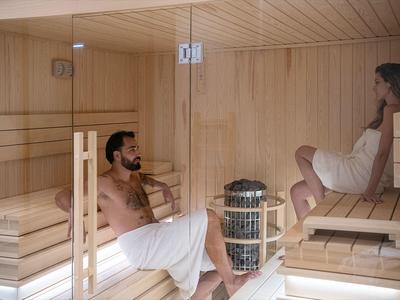 Deux personnes se détendent dans un sauna en bois avec une paroi en verre, toutes deux enveloppées de serviettes blanches.