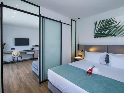 Chambre d'hôtel moderne avec lit double, portes coulissantes en verre et coin salon attenant avec télévision.