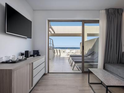 Nowoczesny pokój hotelowy z tarasem i widokiem na morze, przytulny kącik wypoczynkowy i telewizor na ścianie.