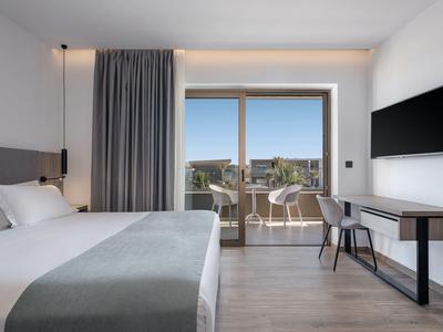 Chambre d'hôtel moderne avec grand lit, bureau, chaises sur balcon et vue sur la ville.