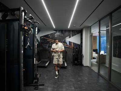 Un uomo cammina in una palestra moderna con pavimento scuro e illuminazione a LED sul soffitto, tenendo un asciugamano.