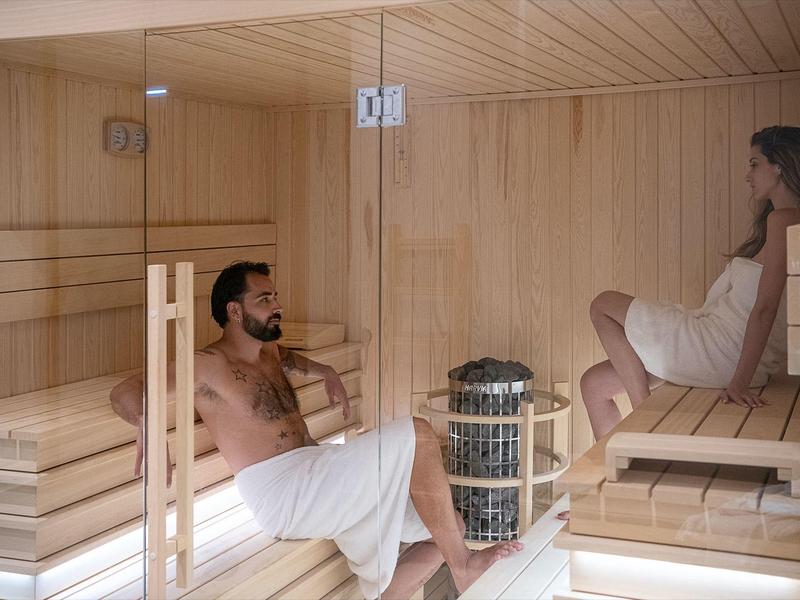 Deux personnes se détendent dans un sauna en bois avec une paroi en verre, toutes deux enveloppées de serviettes blanches.