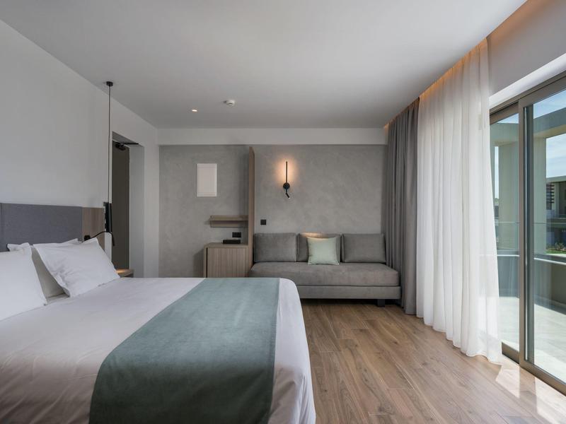 Chambre d'hôtel moderne avec lit double, canapé, sol en bois et balcon avec garde-corps en verre.