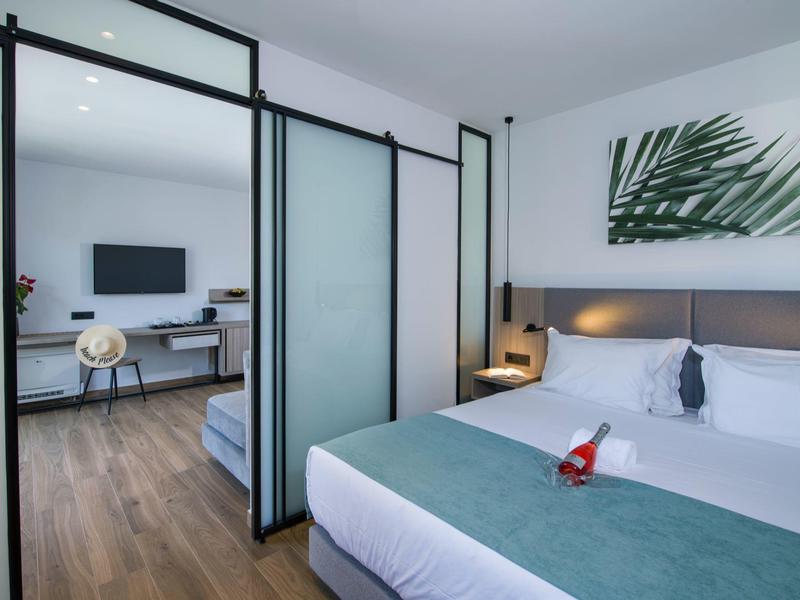 Chambre d'hôtel moderne avec lit double, portes coulissantes en verre et coin salon attenant avec télévision.