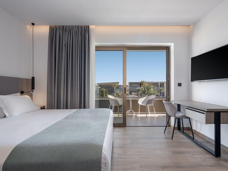 Chambre d'hôtel moderne avec grand lit, bureau, chaises sur balcon et vue sur la ville.