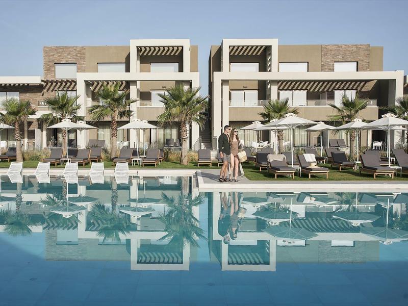 Complexe hôtelier moderne avec grande piscine, palmiers et chaises longues sous un ciel clair.