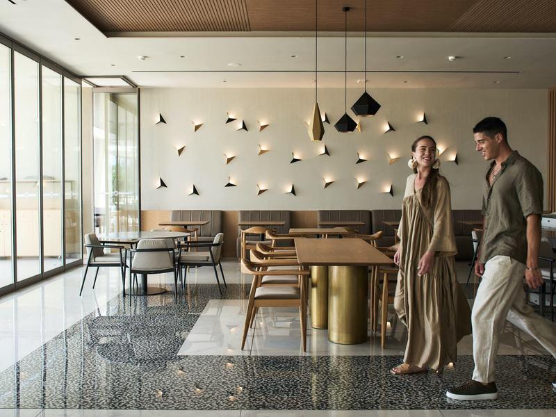 Restaurant d'hôtel moderne avec grandes baies vitrées, mobilier élégant et deux clients qui marchent.