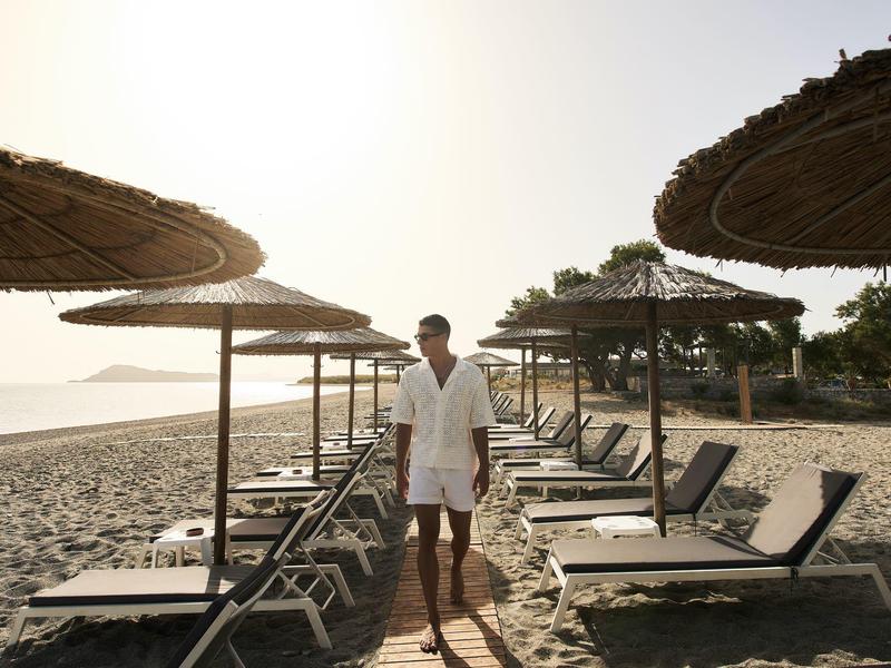 Personne en chemise blanche et short marchant sur la plage entre des transats et parasols.