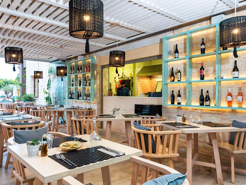 Modern restaurant with wooden tables, chairs, and illuminated wine shelves on the wall.