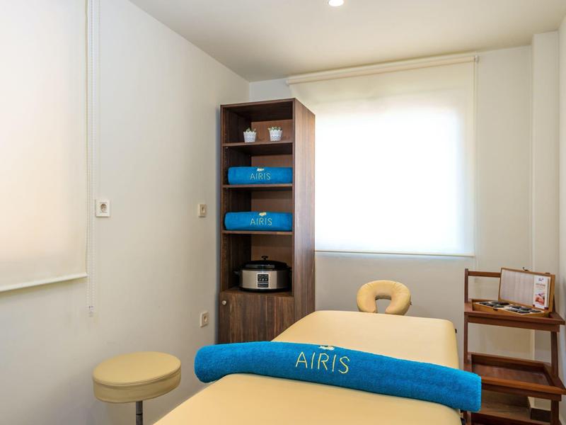 Bright treatment room with massage table, stool, and shelves holding equipment and towels.