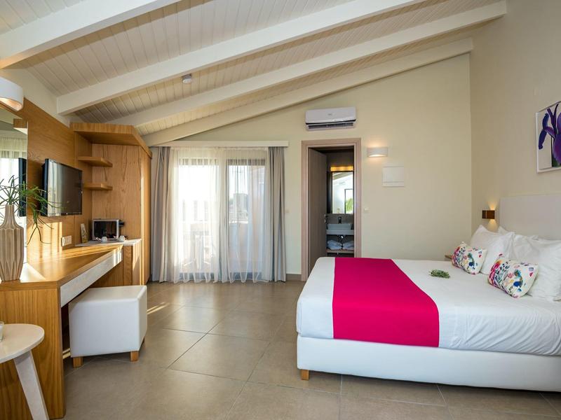 Bright hotel room with double bed, pink bed runner, desk, and TV.