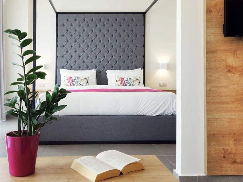 Modern hotel room with a large padded gray headboard, floral-patterned pillows, and a plant in a red pot.