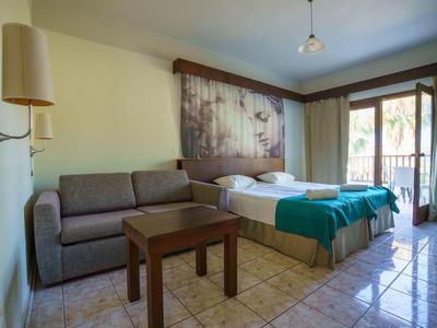 Hotel room with double bed, sofa, wooden table, and balcony overlooking palm trees.