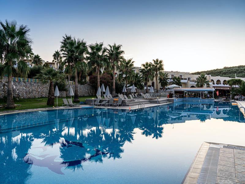 Large pool with palm trees and sun umbrellas in a hotel garden under clear sky.