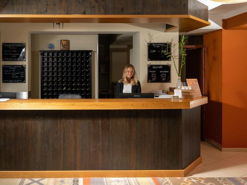 Receptionist sits behind a wooden counter in a warmly lit hotel lobby area.