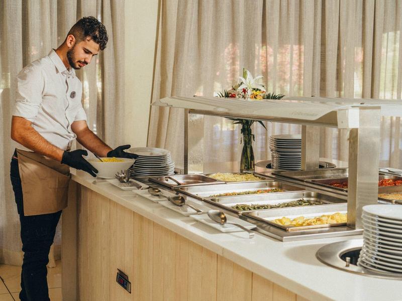 A chef prepares dishes at a buffet counter in a bright room with curtains.