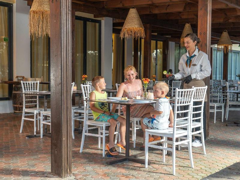 Three children sit at a table in the hotel restaurant as a waitress serves them drinks.