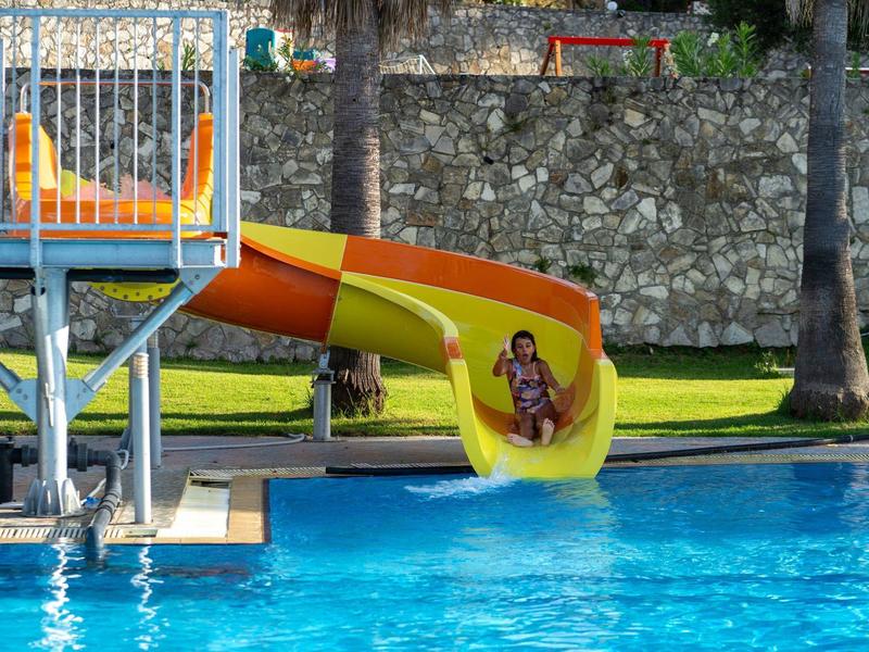 Two children slide down a yellow-orange water slide into a pool with palm trees in the background.