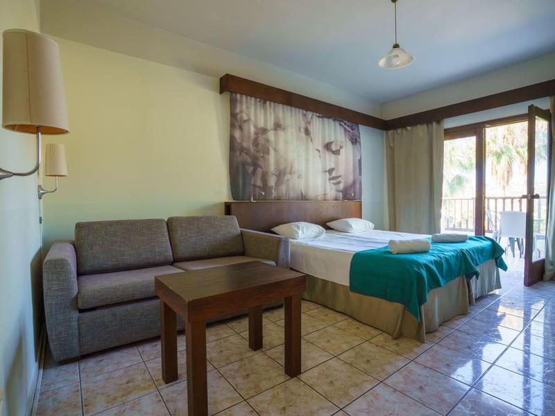 Hotel room with double bed, sofa, wooden table, and balcony overlooking palm trees.