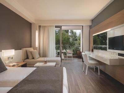 Habitación de hotel moderna con cama, sofá, escritorio, TV y balcón con vistas a árboles.