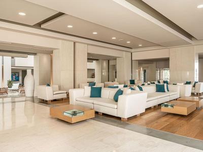 Área de salón luminosa con sofás blancos, cojines azules y mesas de madera en un hotel moderno.