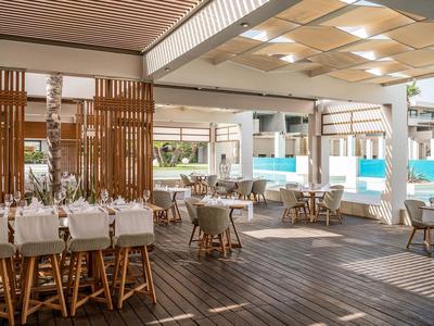 Modernes Hotelrestaurant mit weißen Tischen und Holzstühlen, hellem Holzfußboden und Poolblick.