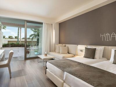 Habitación de hotel luminosa con dos camas, mobiliario moderno y acceso a la terraza con vista a la piscina.