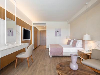 Habitación de hotel moderna con cama doble, TV, escritorio y área de estar en tonos neutros.