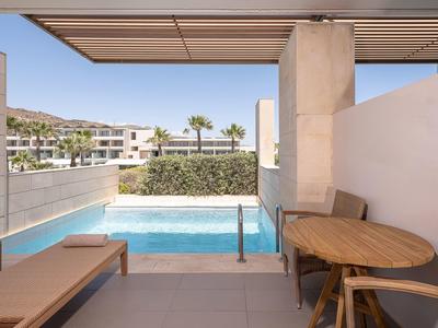 Área exterior moderna con piscina, mesa de madera, banco y vista a palmeras y edificios bajo cielo claro.