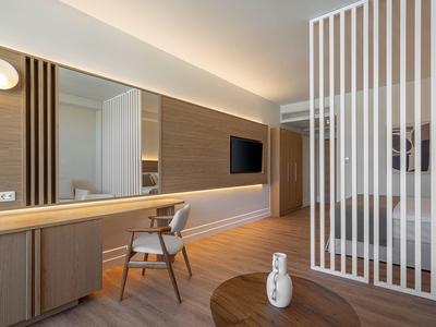 Habitación de hotel modernamente amueblada con escritorio, silla, televisor y separador de ambiente con luz cálida.