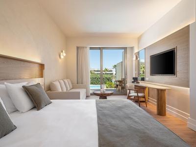 Habitación de hotel luminosa con cama doble cómoda, sofá, escritorio y vista a zona verde exterior.