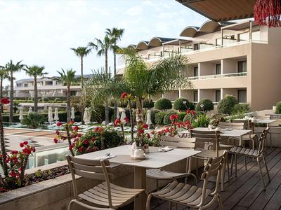 Hotelterrasse mit Holzdeck, Tischen und Stühlen, umgeben von tropischen Pflanzen und Palmen.