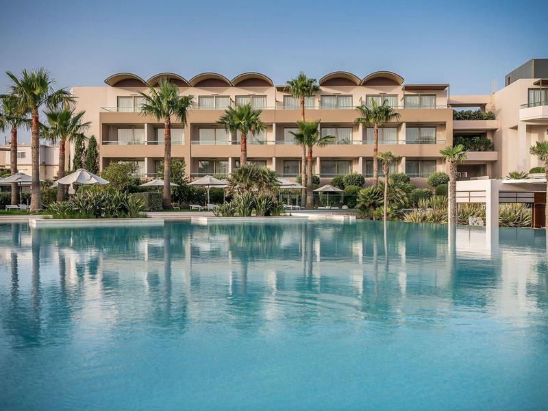 Complejo hotelero moderno con varios pisos, palmeras y gran piscina azul en primer plano.