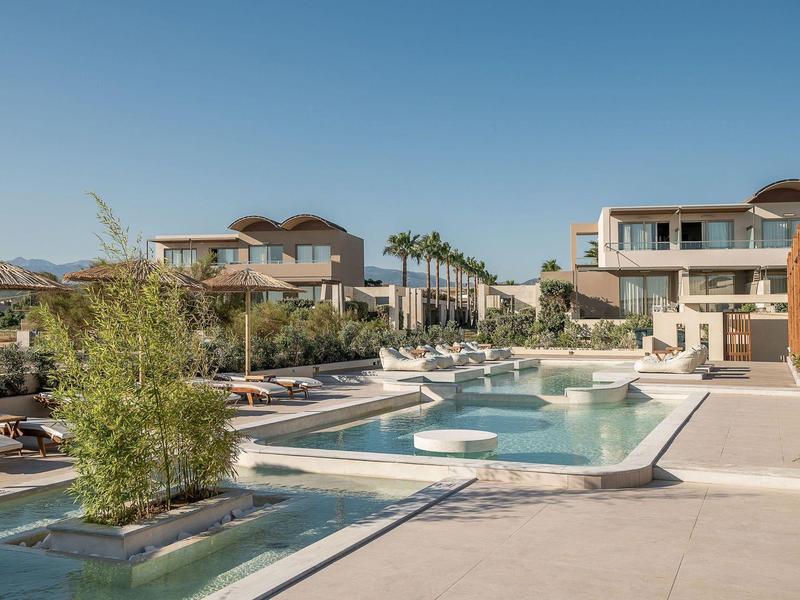 Piscina moderna de hotel con tumbonas, plantas y edificios de varios pisos bajo cielo despejado.