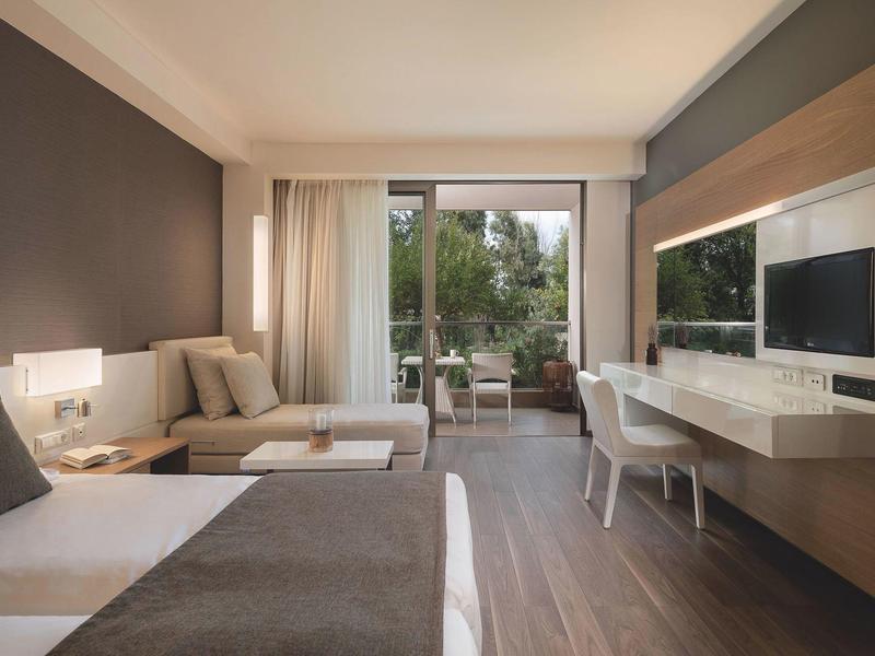 Habitación de hotel moderna con cama, sofá, escritorio, TV y balcón con vistas a árboles.