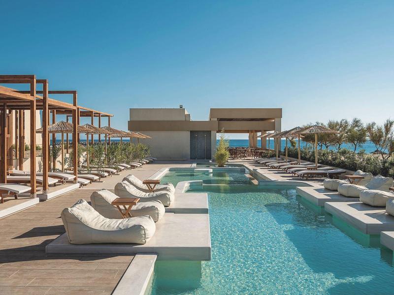 Área moderna de piscina con tumbonas y pabellones bajo un cielo azul en un hotel.