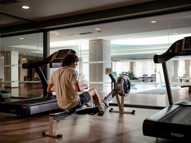 Persona entrenando en una máquina de remo en un gimnasio moderno de hotel con grandes ventanales.