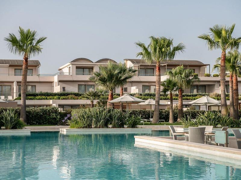 Vista de un hotel con piscina, palmeras y tumbonas en un día soleado.