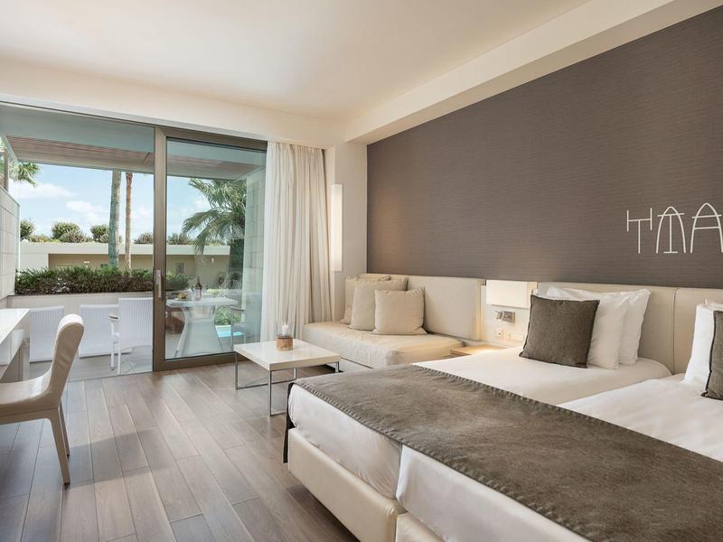 Habitación de hotel luminosa con dos camas, mobiliario moderno y acceso a la terraza con vista a la piscina.