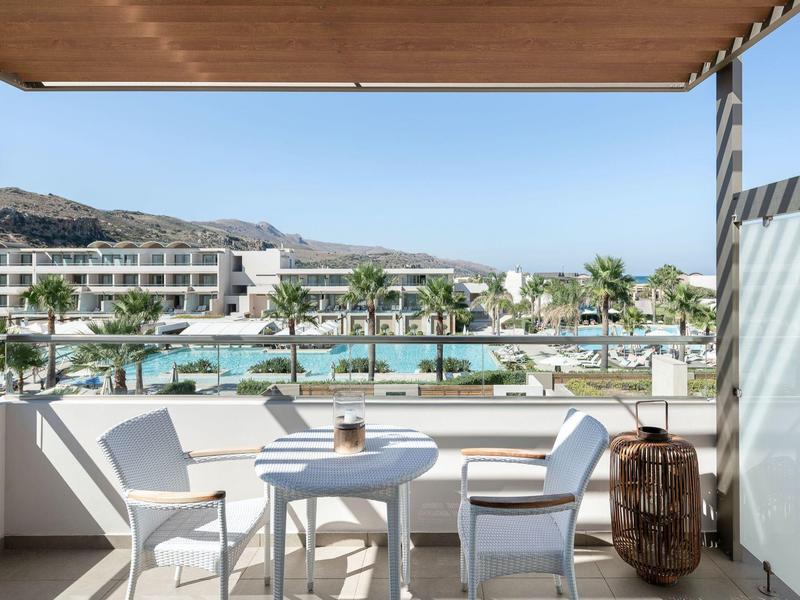 Balcón con mesa y sillas con vista a la piscina y moderno hotel bajo el cielo azul.