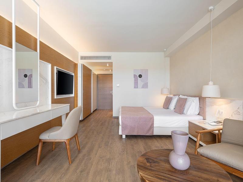 Habitación de hotel moderna con cama doble, TV, escritorio y área de estar en tonos neutros.