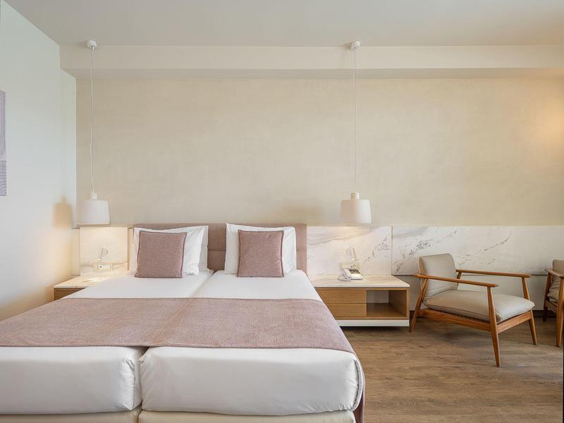 Habitación de hotel moderna con cama doble, dos almohadas, luces nocturnas y un sillón junto a la ventana.