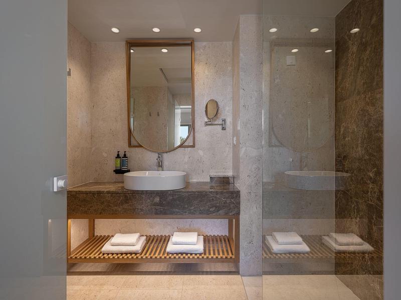 Baño moderno de hotel con lavabo, espejo y toallas sobre un estante de madera.