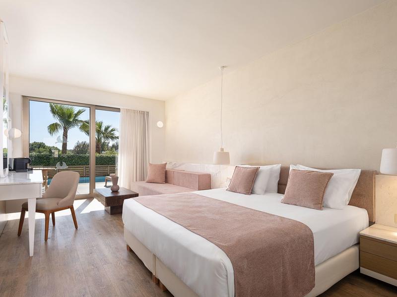 Habitación de hotel luminosa con cama doble, escritorio, silla y vista a palmeras a través de grandes ventanas.