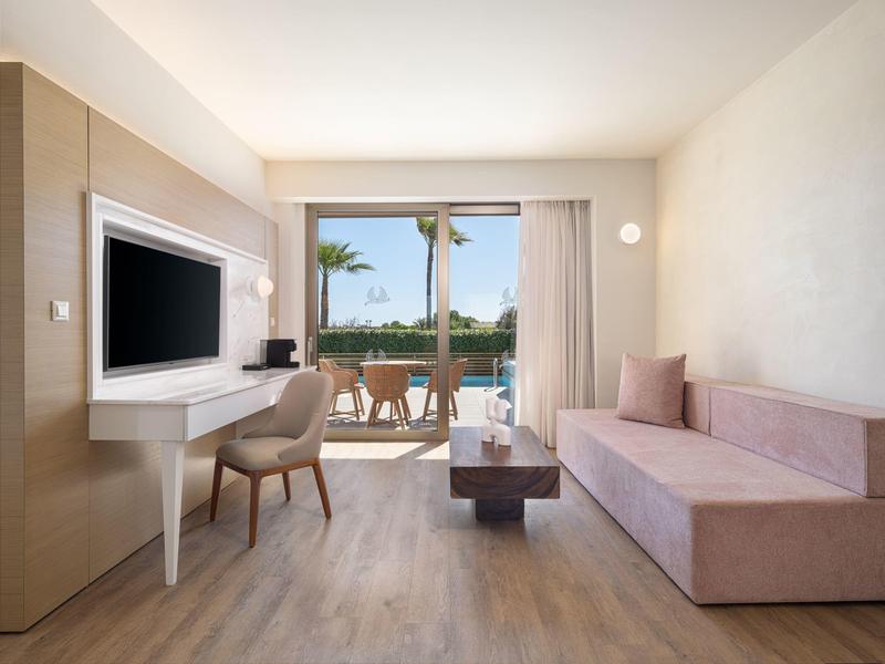 Habitación de hotel luminosa con televisor, escritorio, sofá rosa y balcón con vista a palmeras.