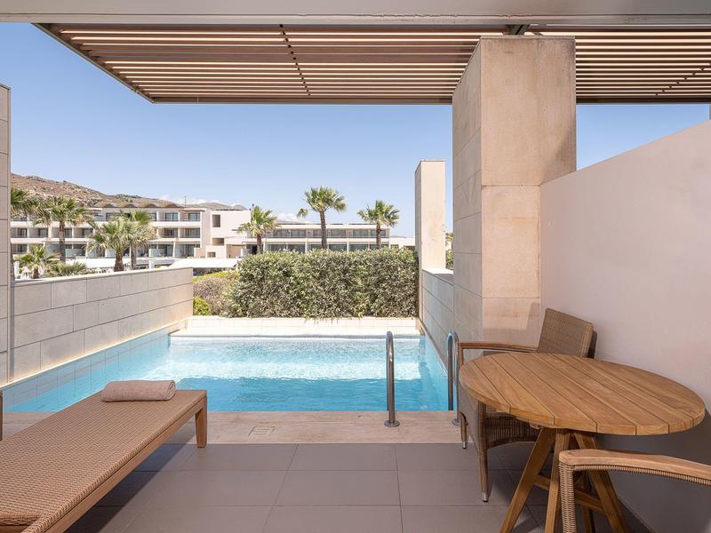 Área exterior moderna con piscina, mesa de madera, banco y vista a palmeras y edificios bajo cielo claro.