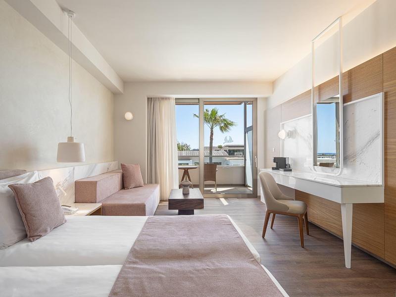 Habitación de hotel luminosa con cama doble, sofá, escritorio y balcón con vista al mar.