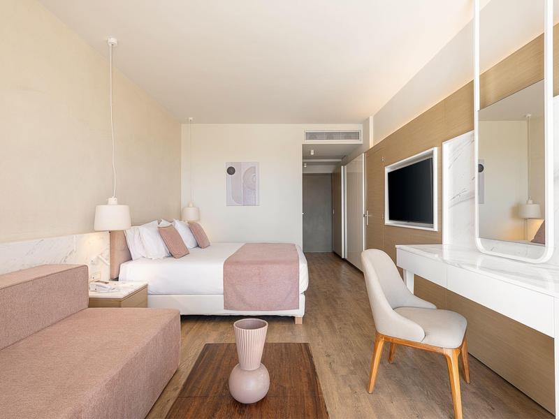 Habitación de hotel moderna y luminosa con cama doble, sofá, escritorio y televisor en la pared.