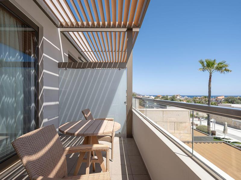 Balcón con silla de madera y mesa, pérgola moderna, vista de palmeras y cielo azul claro.