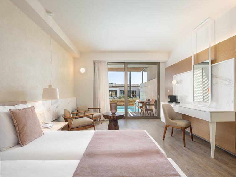 Habitación de hotel luminosa con cama doble, sillas, escritorio y balcón con vista exterior.